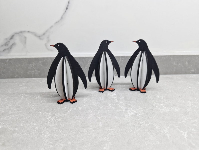 May include: Three black and white penguin figurines standing on a gray surface. The penguins are made of wood and have a 3D design.