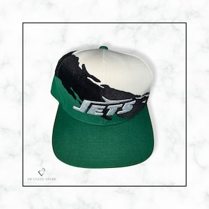 May include: A green, white, and black baseball cap with a white "JETS" logo on the front. The cap has a green brim and a white crown with a black abstract design.