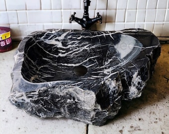May include: A black marble sink with a rough, natural exterior. The sink is oval-shaped and has a white marble vein pattern. The sink is mounted on a white tiled wall.