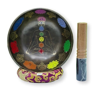 May include: A black singing bowl with a gold design of a meditating figure with seven chakras. The bowl is on a purple cushion and comes with a wooden striker. "Made in Nepal" is printed on the cushion.
