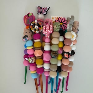 Pode incluir: Um conjunto de agulhas de crochê coloridas com contas e pingentes. Os pingentes incluem um sinal de paz, um cacto, uma flor, uma borboleta e uma maçã com a palavra "Teach" nela.