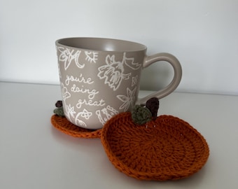 Puede incluir: Una taza de cerámica beige con el texto "you're doing great" en letras blancas. La taza está sobre dos posavasos de calabaza naranja hechos a crochet.