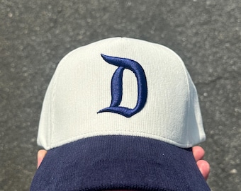 Könnte beinhalten: Eine weiße Baseballkappe mit marineblauem Schirm und einem großen, gestickten marineblauen Buchstaben "D" auf der Vorderseite. Die Kappe ist aus einem strukturierten Stoff gefertigt.