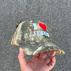 May include: Camouflage baseball cap with the text "I ❤️ JESUS" in white and a red heart. The cap has a curved brim and a tag attached. The hat is held up by a hand.