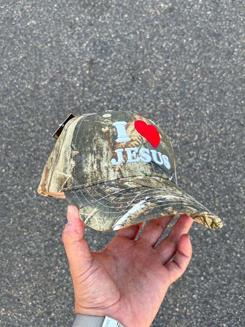 May include: Camouflage baseball cap with the text "I ❤️ JESUS" in white and a red heart. The cap has a curved brim and a tag attached. The hat is held up by a hand.