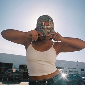 May include: A camouflage baseball cap with the text "I ❤️ JESUS" in white and red. The cap is worn by a person wearing a white tank top and blue jeans. The background includes a building and vehicles.
