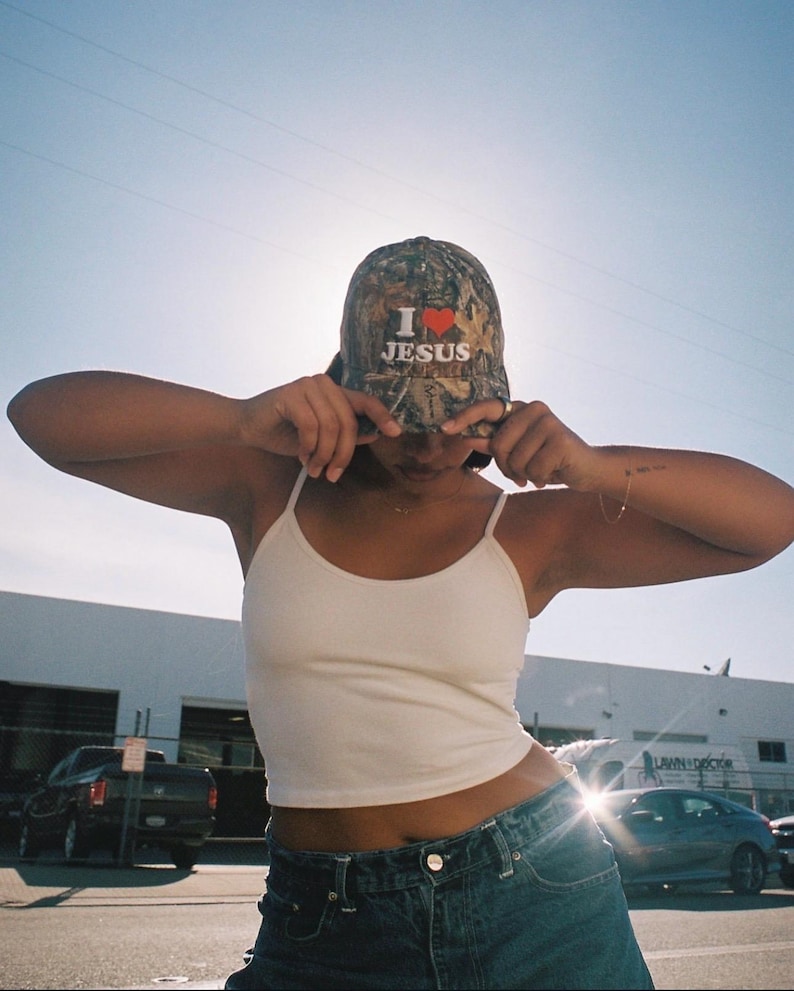 May include: A camouflage baseball cap with the text "I ❤️ JESUS" in white and red. The cap is worn by a person wearing a white tank top and blue jeans. The background includes a building and vehicles.