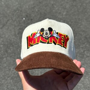 May include: A white and brown corduroy baseball cap with a red and yellow embroidered Mickey Mouse graphic and the word "MICKEY" in red.