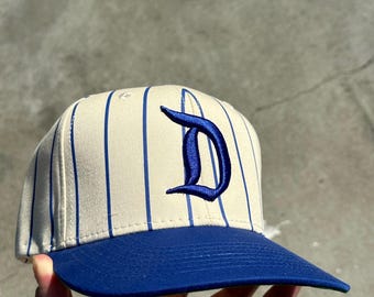 Könnte beinhalten: Baseballkappe mit cremefarbener Krone mit blauen vertikalen Streifen und einem gebogenen blauen Schirm. Die Kappe zeigt einen großen, gestickten blauen Buchstaben "D" auf der Vorderseite. Ein Hut im klassischen Stil, geeignet für Sport oder Freizeit.