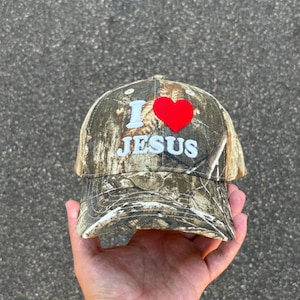 May include: Camouflage baseball cap with the text "I ❤️ JESUS" embroidered in white and a red heart. The cap has a curved brim and a classic six-panel design.
