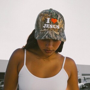 May include: A camouflage baseball cap with the text "I ❤️ JESUS" in white and a red heart. The cap has a curved brim and a structured crown. The background is a soft, neutral tone.
