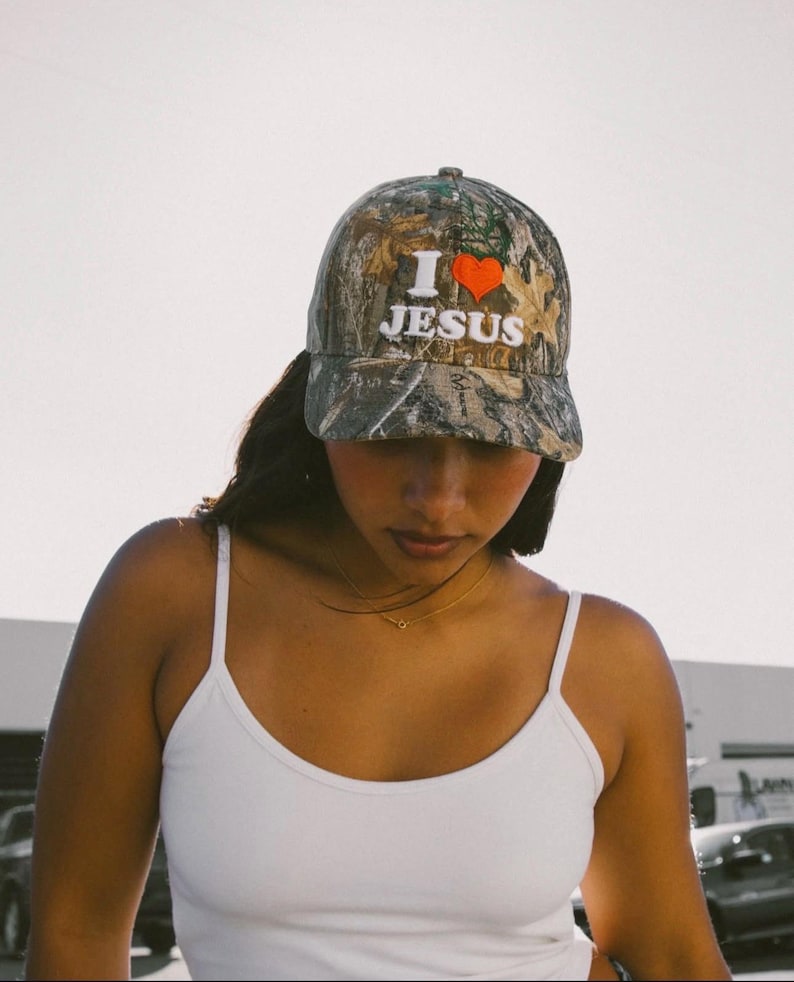 May include: A camouflage baseball cap with the text "I ❤️ JESUS" in white and a red heart. The cap has a curved brim and a structured crown. The background is a soft, neutral tone.