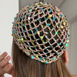May include: A beige crochet hair net featuring an open mesh pattern adorned with numerous small, iridescent, multi-colored sequins in shades of blue, green, pink, and gold. The accessory is circular and covers the crown of the head.