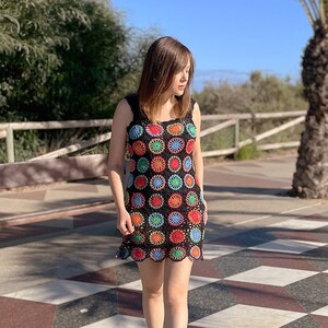 May include: A woman wearing a black crochet dress with colorful circular patterns. The dress has a sleeveless design and a scalloped hem. She is walking on a checkered tiled floor.