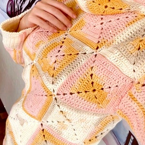 Könnte beinhalten: Ein gehäkelter Pullover mit hohem Kragen und einem geometrischen Muster in den Farben Rosa, Beige und Gelb.