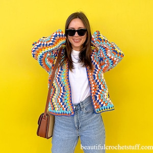 May include: A woman wearing a colourful crocheted cardigan with a white t-shirt and blue jeans. She is standing in front of a yellow wall.