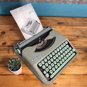 May include: A vintage Hermes Baby typewriter in a light green color, with a matching keyboard and black accents. The typewriter has a manual, and a small succulent plant in a pink pot is next to it. The text "HERMES by" is visible.