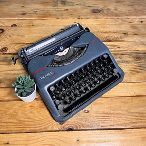 May include: A dark blue Hermes Baby portable typewriter with a QWERTY keyboard layout, black circular keys, and a red accent line across the top casing. The machine features a metal carriage, a ribbon spool cover, and a space bar.