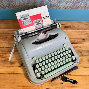 May include: A sage green Hermes Baby portable typewriter with an AZERTY keyboard layout. A manual titled Comment utiliser votre HERMES portative is inserted in the carriage. A black cleaning brush with the Hermes logo is placed nearby.