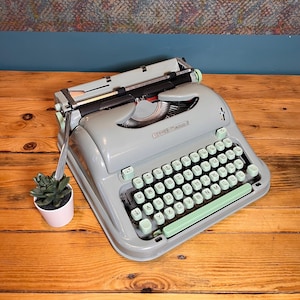 May include: A vintage gray Hermes 3 typewriter with mint green keys and accents. The typewriter sits on a wooden surface next to a small potted succulent. The keyboard features white letter keys.