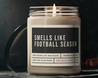 Puede incluir: Una vela de vidrio marrón con una etiqueta blanca que dice "Smells Like Football Season". La etiqueta también incluye el texto "100% Natural Soy Wax Blend", "Burn Time: 50-60 Hours", "Poured in the USA", y "Chuckle Wicks".