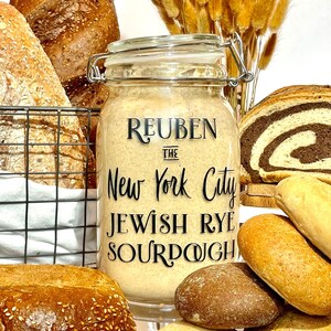 Könnte beinhalten: Ein Glasgefäß gefüllt mit einem hellbraunen Sauerteigstarter mit der Aufschrift "Reuben the New York City Jewish Rye Sourdough". Das Gefäß ist von verschiedenen Broten umgeben, darunter ein dunkles Roggenbrot, ein rundes Brot mit Sesamkörnern und mehrere kleinere Brötchen.