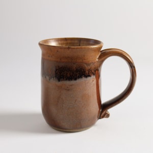 May include: A brown ceramic mug with a dark brown glaze and a light brown rim. The mug has a rounded body and a handle.