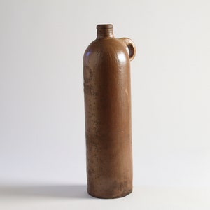 May include: A brown stoneware bottle with a handle and a circular mark on the front. The bottle is about 12 inches tall.