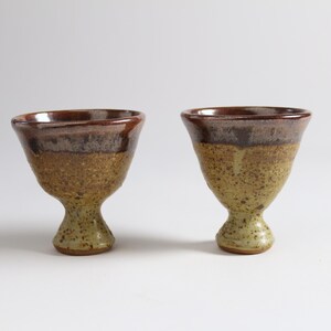 May include: Two brown and yellow ceramic cups with a footed base. The cups have a speckled glaze.