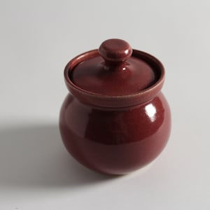 May include: A small, round, red ceramic jar with a lid. The jar has a smooth, glossy finish.