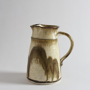 May include: A ceramic pitcher with a cream-colored base and brown drip glaze design. The pitcher has a curved handle and a slightly flared rim. The overall shape is cylindrical, and the base is dark brown.