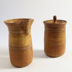May include: A set of two brown ceramic pottery pieces, a pitcher and a jar with a lid. The pitcher has a wide mouth and a slightly curved handle. The jar has a round lid with a knob on top.