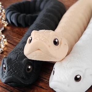 Könnte beinhalten: Drei Spielzeugschlangen, eine schwarze, eine weiße und eine beige, mit realistischen Schlangenmerkmalen, einschließlich Augen und Schuppen.