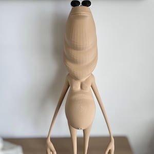 May include: A tan-colored, stylized figurine with a long, oval head and small, round black eyes. The figurine has a slender body, arms, and legs, with a rounded belly. The figure is standing upright with arms outstretched.