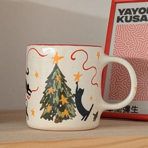 May include: A ceramic mug with a festive design. The off-white mug features a Christmas tree, black cat silhouettes, and star accents. The rim is red, and the handle is white. The mug is on a wooden shelf.