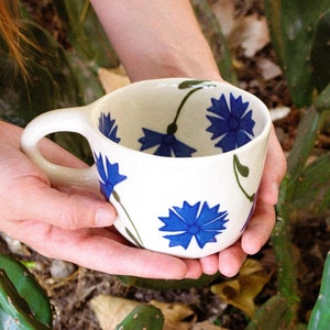 Puede incluir: Una taza de cerámica blanca con un diseño floral azul, sostenida en las manos. La taza tiene una forma redondeada y un asa grande. El fondo incluye plantas de cactus verdes.