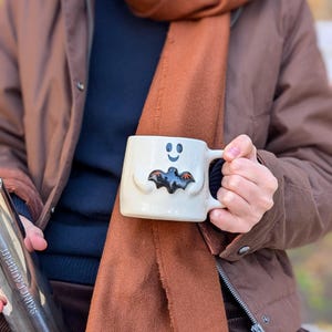 Może przedstawiać: Kremowy kubek ceramiczny z wzorem w kształcie ducha, uśmiechniętą buzią oraz małą, czarną, trójwymiarową figurką nietoperza przymocowaną z przodu. Kubek posiada uchwyt po prawej stronie oraz gładkie, błyszczące wykończenie powierzchni.