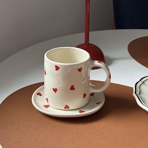 Op de afbeelding: Een witte keramische mok en schotel set versierd met kleine rode hartjes. De mok staat op een bijpassende schotel, geplaatst op een bruine placemat. Een rode tafellamp met een ronde kap staat op de achtergrond.