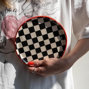 May include: A round ceramic plate with a black and white chequerboard pattern and a red rim. The plate is held in front of a white t-shirt with a graphic design.