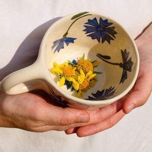 Puede incluir: Un cuenco de cerámica color crema con un asa, decorado con motivos florales azules. El cuenco contiene un pequeño ramo de flores amarillas. El cuenco se sostiene con dos manos.