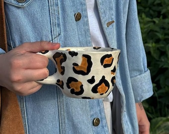 May include: A white ceramic mug with a leopard print design in black and brown. The mug has a large handle and is being held by a person wearing a denim jacket and carrying a brown suede bag.