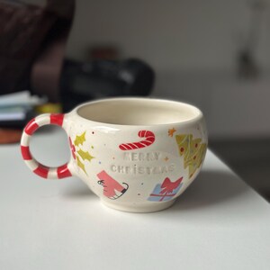 Op de afbeelding: Crèmekleurige keramische mok met een rood-wit gestreept oor. De mok is versierd met kerstthema-illustraties, waaronder een zuurstok, kerstboom en de tekst "MERRY CHRISTMAS".