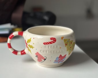 May include: Cream-colored ceramic mug with a red and white striped handle. The mug is decorated with Christmas-themed illustrations, including a candy cane, Christmas tree, and the words "MERRY CHRISTMAS".