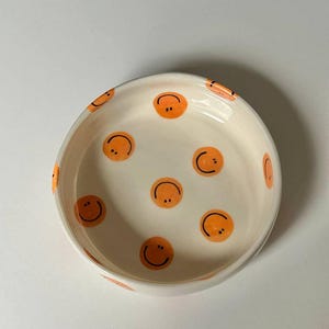 Puede incluir: Un cuenco de cerámica blanco con caras sonrientes naranjas. El cuenco tiene forma redondeada y una superficie lisa. Las caras sonrientes están distribuidas por el interior del cuenco.