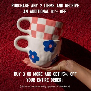 May include: Two ceramic mugs stacked on top of each other, held by a hand, against a red background. The top mug has a pink and white checkered pattern, while the bottom mug is white with blue flower designs. Text on the image reads: "PURCHASE ANY 2 ITEMS AND RECEIVE AN ADDITIONAL 10% OFF! BUY 3 OR MORE AND GET 15% OFF YOUR ENTIRE ORDER!"