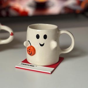 May include: A cream-colored ceramic mug with a ghost face and a pumpkin decoration. The mug sits on a red Pantone coaster with white text. The mug has a handle and two small arms.