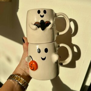 May include: Two stacked white ceramic Halloween mugs. The top mug has a black bat design, and the bottom mug has an orange pumpkin design. Both mugs have black dot eyes and a curved smile.