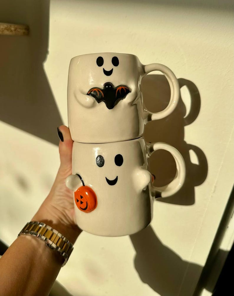 May include: Two stacked white ceramic Halloween mugs. The top mug has a black bat design, and the bottom mug has an orange pumpkin design. Both mugs have black dot eyes and a curved smile.
