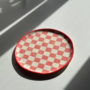 May include: A red and white checked ceramic plate with a red rim. The plate is round and has a glossy finish.