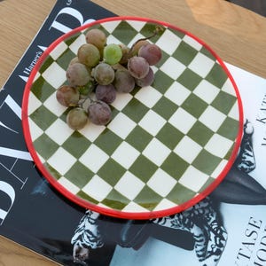 May include: A decorative plate with a green and white checkered pattern and a red rim, holding a bunch of grapes. The plate rests on a magazine with the word "Harper's" visible.